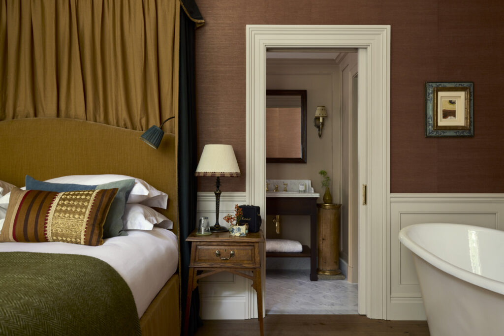 A cosy bedroom with a mustard headboard, layered pillows, wooden bedside table with a lamp, and an open doorway leading to a bathroom with a mirror, lamp, and bath.